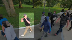A highlighted still from a Nationalist Social Club propaganda video, depicting their daytime march through Boston Common during July 2021. Nine members march in roughly two stacks. Hood (highlighted) in his preferred red Oakley Holbrook sunglasses, a slate baseball cap, black polo shirt, and khakis, trails behind two other members, with the other six trailing behind him.