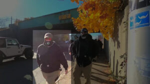 A highlighted still from a Nationalist Social Club propaganda video, depicting their daytime march toward the Lucy Parsons Center in Jamaica Plain MA during November 2021. In the foreground, from left to right, are Hood (highlighted) and Leo Cullinan walking along a sidewalk. Hood is wearing in his preferred red Oakley Holbrook sunglasses, a black gaiter with a stylized white "H" over the mouth, a slate baseball cap with an unidentifiable patch, a black Carhartt hoodie, and khakis. In the background, several members of NSC follow.