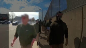 A highlighted still from a Nationalist Social Club propaganda video, depicting their daytime march toward Boston's Saint Patrick's Day parade during March 2022. In the foreground, from left to right, are Hood (highlighted) and an unidentified NSC member as they march along the sidewalk. Hood wears a baseball cap, his preferred red Oakley Holbrook sunglasses, and an olive green Champion hoodie. Hood' face is blurred, but the Oakleys are still readily identifiable. In the background is a procession of NSC members, walking in two stacks.