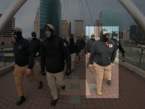 A highlighted still from a Nationalist Social Club propaganda video, depicting their daytime march through Hartford CT during April 2022. In the foreground, from left to right, are Chase Gilroy, an unidentified NSC member, and Hood (highlighted) and an unidentified NSC member as they cross a bridge. Hood wears a Condor Tactical baseball cap with a "131" patch on the front, a black gaiter with a stylized white "H" over the mouth, a black Carhartt hoodie personalized with "131" in white lettering on the left breast, his preferred red Oakley Holbrook sunglasses hanging from the hoodie's collar, and khakis. In the background are several other NSC members.