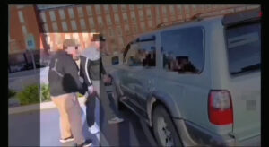 A highlighted still from a Nationalist Social Club propaganda video, depicting their daytime stalking of a local activist in Manchester NH during May Day 2022. From left to right are Hood (highlighted), Leo Cullinan, and the local activist's vehicle. Both Hood and Cullinan are standing facing the driver's side window of the car, and their faces are pixelized to ostensibly disguise their identities. Hood wears a olive or brown baseball cap, a black Carhartt hoodie, a bodycam chest harness, and khakis.