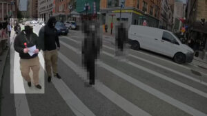 A highlighted still from a Nationalist Social Club propaganda video, depicting their daytime standout outside of Park Street Station in Boston MA during April 2022. In the foreground, from left to right, are Hood (highlighted), Leo Cullinan, and two passers-by occluded by pixelization. Hood wears a tan Condor Tactical baseball cap with a "131" patch on the front, a black gaiter with a stylized white "H" over the mouth, a black hoodie, a bodycam chest harness, his preferred red Oakley Holbrook sunglasses hanging from the harness, and khakis. Hood is also holding a piece of paper, likely a propaganda flyer. Cullinan is positioned to screen Hood from the passers-by.
