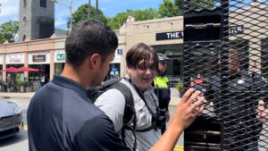 A still from video recorded by Rod Webber, depicting Chris Hood's arrest by BPD in Jamaica Plain MA, after the Nationalist Social Club's daytime protest of a Drag Queen Story Hour during July 2022. In the immediate foreground is an unidentified BPD officer, holding open the back door to a police wagon. In the mid-ground, framed on the left by the officer in the foreground, and on the right by the wagon's door, is Hood. Hood wears a white button-up shirt with pockets on both breasts, a black backpack, and a bodycam chest harness. He is unmasked, and grinning at the camera, possibly in disbelief. Hood's hair is a greasy mess, likely from basting in his own sweat under his baseball cap, in the sun, all afternoon.