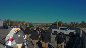 A highlighted still from a Nationalist Social Club propaganda video, depicting their daytime march through Kingston MA during October 2022. In the immediate foreground, to the left of frame, is Hood (highlighted) as he leads several NSC members, including Leo Cullinan and unidentified member designation "Longneck," through a parking lot. Hood wears a tan Condor Tactical baseball cap with a "131" patch on the front, a black gaiter with a stylized white "H" over the mouth, his preferred red Oakley Holbrook sunglasses, a black Carhartt hoodie personalized with "131" in white lettering on the left breast, and khakis. He carries a red and white bullhorn in his right hand, and has a black walkie-talkie with teal trim clipped to a pocket on his pants leg.