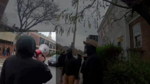 A highlighted still from a Nationalist Social Club propaganda video, depicting their overcast daytime attempt to disrupt the Boston Anarchist Bookfair at the Democracy Center in Cambridge MA during November 2022. In the immediate foreground, to the left of frame, is Hood (highlighted) as he squawks on his bullhorn. Hood wears a black hoodie with the hood up, and a tan baseball cap that peeks out of the hood. Center of frame are a number of NSC members, milling about the sidewalk in front of the Democracy Center. To the right of frame, in the background, is the Democracy Center. Largely barren trees are overhead.