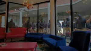 A highlighted still from a video shot by drag performer Juicy Garland from within the Teatotaller, as the Nationalist Social Club lined up outside the building in an attempt to disrupt the day's Drag Queen Story Hour in Concord NH during June 2023. The inside of the Teatotaller is visually warm and comfortable, with an array of colorful couches and a small chandelier overhead. Flower beds line the room's large exterior windows. Outside those windows are a visible line of NSC members, including Farrea and Gilroy, in the middle of a chant. Passing behind the NSC members, barely visible between two unidentified members, is Hood (highlighted). Hood is wearing a tan baseball cap, his Oakley Jawbreakers, some variety of dark-colored shirt, a black backpack, and khakis. He's also holding some manner of selfie stick, at the far end of which a video camera of some kind is attached.
