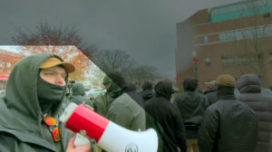 A highlighted still from a Nationalist Social Club propaganda video, depicting their overcast daytime attempt to disrupt a Drag Queen Story Hour held at the People's University Public Library in Fall River MA during December 2022. In the immediate foreground, to the lower left of frame, is Hood (highlighted) as he squawks on his red-and-white bullhorn. Hood wears a tan Carhartt baseball cap, a black gaiter, a black hoodie with the hood up, and his preferred red Oakley Holbrooks hanging from the hoodie's collar. The gaiter is partially sucked into his mouth, as he breathes in to continue blathering on his bullhorn. Lower center and lower right of frame are a number of NSC members, including Cullinan and designation "Longneck," mostly standing in a row with their backs to the library and the point-of-view. In the background, to the upper right of frame, is the 79 North Main Street building, a brutalist-style office complex largely built of brick. In the background, to the upper left of frame are a number of distant trees, either barren or still bearing red leaves from the fall.