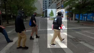 A highlighted still from a Nationalist Social Club propaganda video, depicting their march through Boston MA during their Holocaust Memorial standout during May 2021. In center frame is Hood (highlighted) as he leads members of NSC, including Tyler Moody, along a crosswalk. Hood wears an off-black Columbia baseball cap, a black North-Face-branded long-sleeve that almost looks like a screenprinted knock-off, khakis, and black sneakers with white soles. Slung over his right shoulder is a carrying strap that holds his red-and-white bullhorn. This is a side-view of Hood that occludes his face, but demonstrates an identifying element of his gait: Hood stands and walks with a forward cant at his hips. This suggests tight hip flexor muscles, possibly due to a lower back injury or a weak posterior chain.