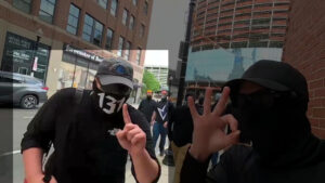 A highlighted still from a Nationalist Social Club propaganda video, depicting their march through Boston MA during their Holocaust Memorial standout during May 2021. In the immediate foreground, from left to right, is Hood (highlighted) and an unidentified NSC member as they lead other NSC members in the background, including Liam MacNeil, through the streets of Boston. Hood wears an off-black Columbia baseball cap with a stylized mountain range or trio of trees as a front emblem, black sunglasses that bear some resemblance to Oakley Jawbreakers, a black gaiter with "131" in white lettering across the face, and a black North-Face-branded long-sleeve shirt with sleeves rolled up to the elbows. Slung over his right shoulder is a carrying strap that holds his red-and-white bullhorn. Hood holds his left hand up with the index finger extended, likely harkening to Nick Fuentes's "America First" movement. The unidentified member next to him, clad in black baseball hat, sunglasses, gaiter, and shirt, holds his right hand in the "okay" gesture.