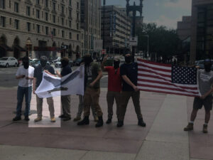 A highlighted picture from a series of photos of Patriot Front's attempts to disrupt the Boston Women's March in August 2019, taken by Emily Goldhammer. Eight members stand in a row, some holding flags. The second, the highlighted member, and the fourth from the left hold a flag of an eagle with spread wings clutching a set of fasces in its talons, seeming to be the war flag symbol used by the Italian Social Republic. The first and second from the right hold an American flag, albeit backwards. From left to right: an unknown Patriot From member, Chris Hood (highlighted), Matthias Thorpe, an unknown Patriot From member, Robert Budzyna, an unknown Patriot From member, Noah Bogosh, and Mark Hayden.