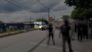 A still from an antifascist's video of members of the Nationalist Social Club fleeing in vehicles after getting rumbled before a daytime vandalization campaign in Manchester NH during August NH. In the foreground to right of frame, as well as the background to left of frame, are several antifascists, pixelized to conceal identities. In the background, center frame, is a white Jeep Wrangler backing out of parking space. The passenger side window is rolled down, and Hood's arm (highlighted) is barely visible deploying pepper spray at a nearby antifascist.