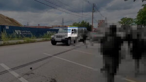 A highlighted still from an antifascist's video of members of the Nationalist Social Club fleeing in vehicles after getting rumbled before a daytime vandalization campaign in Manchester NH during August NH. In the foreground to right of frame, as well as the background to left of frame, are several antifascists, pixelized to conceal identities. In the background, center frame, is a white Jeep Wrangler driving forward. The driver's side window is rolled down, and Hood's arm (highlighted) is barely visible deploying pepper spray at a nearby antifascist just outside the driver's side door, moments before the driver rolls the window up.
