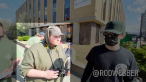 A highlighted still from video recorded by Rod Webber, depicting members of the Nationalist Social Club, as their civic group PINE, in the midst of handing out PINE literature at a daytime Trump rally in Manchester NH during April 2023. In the foreground, from left to right, is Hood (highlighted) and an unidentified NSC member in all black. Hood wears an olive green Carhartt baseball cap, an olive green hoodie with the sleeves rolled to the elbows and the hood up, and a Nikon DSLR camera slung around his neck. He is unmasked, and avoiding eye contact with the point-of view. In the background is the Consolidated Communications building on Elm Street.