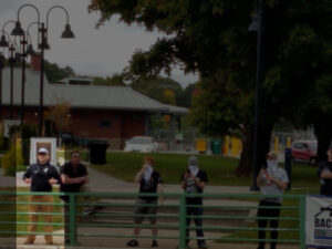 A highlighted photo taken from a distance by an antifascist activist of the Nationalist Social Club, having joined a daytime demonstration by America Backs the Blue in Mansfield MA during September 2020. From left to right, standing in a group behind a set of metal sidewalk barricades, are Hood (highlighted), Anthony Petruccelli, Cameron Anthony, an unidentified NSC member, and Tyler Moody. Every member faces counter-protesters across the street, from which side the photo was taken. Hood wears a gray baseball cap, black shades, what appears to be a long-sleeve black t-shirt with the NSC logo in white on the left breast, a bodycam chest harness, a silver wristwatch on his left, khakis, and work boots.