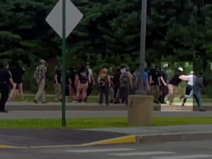 A highlighted still from a Nationalist Social Club propaganda video, shot from a very far distance, depicting counter-protesters placing their bodies between NSC and their staging location across from the Nashua Board of Education building during the daytime in August 2021. Hood (highlighted) can be made out shoving a counter-protester, in an attempt to make way. Hood is visibly wearing a black t-shirt, tan baseball cap, black gaiter, and khakis.