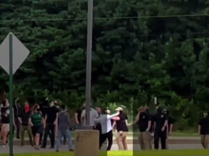 A highlighted still from a Nationalist Social Club propaganda video, shot from a very far distance, depicting counter-protesters placing their bodies between NSC and their staging location across from the Nashua Board of Education building during the daytime in August 2021. Hood (highlighted) can be made out shoving the same counter-protester from the previous still, having progressed further toward the building. Hood is visibly wearing a black t-shirt, bodycam chest harness, tan baseball cap, black gaiter, and khakis.