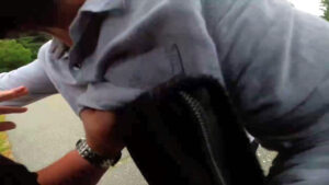 A still from a Nationalist Social Club propaganda video, shot from Hood's bodycam, depicting a shoving match with a counter-protester in Nashua NH during the daytime in August 2021. While Hood is largely not visible, his silver wristwatch is easily visible on his left wrist, as he clutches the counter-protester's shirt. Hood also carries some sort of black, zippered binder in his right hand, which he uses as a surface with which to shove the counter-protester.