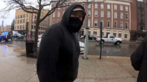 A still from a Nationalist Social Club propaganda video, depicting their overcast daytime protest outside of Portland City Hall in Portland ME during April 2023. This is shortly after their assault of local antifascists and concerned citizens, whereupon the local police held NSC at gunpoint before letting them leave with no members under arrest. In the immediate foreground, center frame, from the waist up, is Hood. He wears a tan Carhartt baseball cap, a black gaiter, and a black Carhartt hoodie with the hood up. There is a faint look of incredulousness on his face. In the background is the part of Portland across from City Hall. To the left, to Hood's back, is a police truck, lights flashing.