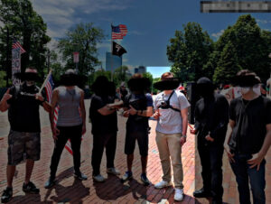 A highlighted picture from a Nationalist Social Club propaganda photo set, depicting a daytime standout at Boston Common during May 2020. From left to right, standing in a row, are Zach Brackett, two unidentified NSC members, Cameron Anthony, Hood (highlighted), Michael Moura, and an unidentified NSC member. Hood's hair is styled into an undercut. He wears a light gray t-shirt with what appears to be an American flag on the left breast pocket, a bodycam chest harness, a silver wristwatch on his left wrist, khakis, and white sneakers. Hood, Brackett, and Moura hold the index fingers of their right hands raised, in a hand gesture that is likely a reference to Nick Fuentes's "American First" movement.