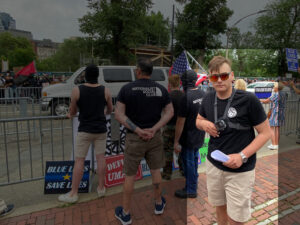 A highlighted photo taken by Gregg Housh of a Nationalist Social Club outside of the Massachusetts State House, having joined a daytime protest of coronavirus lockdown conditions by Super Happy Fun America during June 2020. From left to right, standing in a group at a set of metal police barricades, are Edward Stuart, Anthony Petruccelli, Cameron Anthony, Michael Moura, and Hood (highlighted). The other NSC members face away from the State House, their focus on counter-protesters across the street. Only Hood is looking at the camera. Hood's hair is styled into an undercut. He wears his preferred red Oakley Holbrook sunglasses, a black t-shirt with a white NSC logo on the right breast, a bodycam chest harness, a silver wristwatch on his left wrist, khaki shorts, and white sneakers. Petruccelli and Moura both wear black t-shirts with North Face knock-off logos, with "The North Face" replaced by "Nationalist Social Club" and the numbers "131" in each third of the North Face triple-arch logo. Visibly held by Stuart and Anthony, resting on the barricade, is a white flag with a black sonnenrad on it. At their feet lie several protest signs, likely brought by Super Happy Fun America, including "Blue Lives Save Lives" and "Defund UMass."