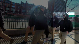 A highlighted still from a Nationalist Social Club propaganda video, depicting NSC walking along the Dr. Paul Dudley White Bike Path during the daytime after their Storrow Drive standout during February 2022. In the foreground are a number of NSC members marching in double-stack. Toward the rear of the march, from left to right, are Leo Cullinan and Hood (highlighted). Hood wears a tan Condor Tactical baseball cap with a "131" patch on the front, what appear to be black Oakley Jawbreaker sunglasses, his stylized "H" gaiter, a Carhartt hoodie, khakis, and black sneakers with white soles. In the background is Storrow Drive; further beyond are several buildings along Back Street.