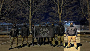 A highlighted propaganda photo taken by a member of the Nationalist Social Club during their nighttime counter-flyering of Liam MacNeil's neighborhood in Waltham MA during August 2022. In the foreground, eight NSC members stand in a row in a parking lot, with the four in the center holding an NSC anticommunist/anticapitalist flag. The outer four members are carrying expandable batons. Second from the right is Leo Cullinan, and first on the right is Hood (highlighted). Hood wears a black gaiter, a black North Face overshell jacket with the hood up, a bodycam chest harness, khakis, and black sneakers with white soles. He holds the baton in his right hand, and has his left tucked into his pants pocket. In the background is a chainlink fence in front of bare trees, Further still appears to be the Charles River, with more cityscape across the water.