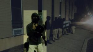 The Atlantic Nationalist Club, staging an after-hours protest outside of the One People's Project Office in New Brunswick NJ during December 2024. Members largely wear black hoodies or long-sleeved t-shirts, pale khaki pants, black baseball caps, black footwear, and black facial coverings. One member appears to be Charles Netter. He wears a black long-sleeve hoodie with the hood up, brown work gloves, and a black ski mask. He speaks into a black megaphone. Several members stand in a line behind him on the sidewalk, with a number of them holding a banner redacted by the editor.