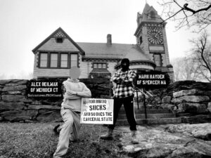 A monochrome photo of two men posing with a law sign in front of the Princeton Public Library in Princeton MA. The lawn sign has been intentionally defaced to say "Rob Rundo SUCKS and so does the carceral state." The man kneeling to the left of the sign has been marked "Alex Beilman of Meriden CT (he's not even FROM here)." The man standing to the right of the sign has been marked "Mark Hayden of Spencer MA." Both men have their arms crossed.