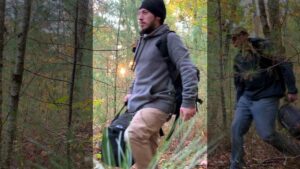 An image of Cerce hiking with other Patriot Front members toward Purgatory Chasm in Sutton MA during October 2021. Cerce, wearing a black beanie, gray hoodie, khakis, and a black backpack, carries a black duffle as he walks through underbrush. He is followed by Patriot Front member Alex Beilman.