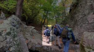 An image of Cerce hiking with other Patriot Front members through Purgatory Chasm in Sutton MA during October 2021. Cerce, wearing a black beanie, gray hoodie, khakis, and a black backpack with a green patch on the back, carries a black duffle as he hikes up rocky terrain.