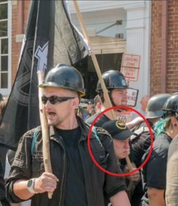 An image of Cerce during Unite the Right, marching with several other members of the Traditionalist Workers Party. He is peeking out from between two other, much taller members. He wears a black baseball cap with a yellow "Anti-Communist Action" emblem on the front.