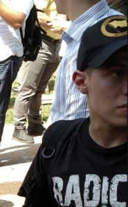 An image of Cerce during Unite the Right. He wears a black baseball cap with a yellow "Anti-Communist Action" emblem on the front, and a black t-shirt with the word "Radic" on the front (likely meant to spell out "Radical Agenda").