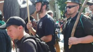 An image of Cerce during Unite the Right, marching with several other members of the Traditionalist Workers Party. He is standing foreground to two other, much taller members. He wears a black baseball cap with a yellow "Anti-Communist Action" emblem on the front, and a black backpack.