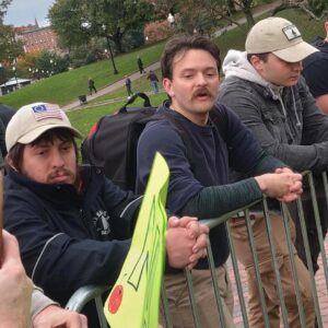 A photo of three members of Patriot Front, leaning forward against a metal barricade in Boston Common during the Men's March of November 2023. From left to right is Cerce, Brian Harwood of Spencer MA, and an unidentified member. Cerce is dressed in a navy blue jacket with a "Bar Harbor ME" emblem on the breast, a tan baseball cap with a Betsy Ross flag patch on the front, and khakis. He leans forward against a metal barricade, which stands at a height between his elbows and shoulders.