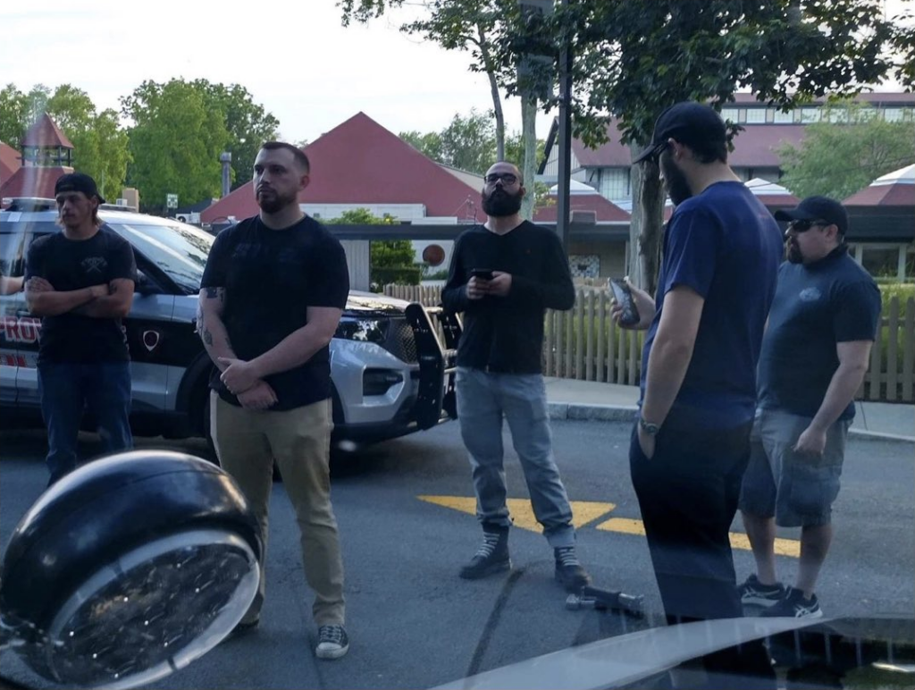 A photo of five members of NSC-131 at a police stop in East Providence RI during June 2022. From left to right are Cody Baker, Austin Conti, Tyler Moody, Stephen Farrea, and Jason Lowe. They are dressed in normal street clothes that give no obvious indication of their NSC membership, save Moody's boots: the laces are white and straight-laced. Both Moody and Farrea are on their phones.