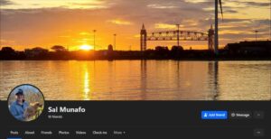 A screenshot of known Patriot Front member Sal Munafo's Facebook profile, specifically the top bar. The top banner image depicts a horizon during sunset in Buzzards Bay MA. Specifically, it is taken somewhere in the water to the west of Taylor Point. Across the water is a landmass, with a wind turbine in the far right midground, the Cape Cod Canal Railroad Bridge in the right background, and the Bourne Bridge partially distorted by the setting sun in the left-of-center distant background. In his profile photo, a grinning Munafo wears a cornflower blue windbreaker and a dark blue Columbia-branded baseball cap, and holds a chicken.