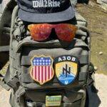 A photo of a tactical backpack. On it are a number of neo-Nazi apparel and patches, including a Will2Rise baseball cap, an Azov battalion patch, and a pair of red mirrored Hammershades.