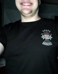 A photo. It is a close-up of an early-20s white man wearing a black t-shirt, indoors. The shirt is a Will2Rise product, with a skeletonized hand grasping five arrows. His face below the nose is visible.