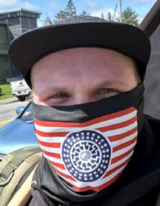 A photo. It is an extreme close-up of an early-20s white man wearing a baseball cap and a gaiter, outdoors. The gaiter is decorated with an American-flag-themed sonnenrad. His eyes and forehead are visible.