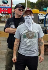 A photo of Ryan Irwin and someone who appears to be NSC-131 co-founder Zach Brackett, at an America Backs the Blue event in Mansfield MA during September 2020. They stand in the northeast corner of the lot for the Mansfield Gulf and Service Station on Chauncy Street, with "The Pro's Auto Care Complex" visible in the background. Irwin wears black pants, a gray Smokey the Bear "If Not You, Who?" graphic t-shirt, black plastic-framed rectangular lenses, and a camo Red Sox baseball cap. He wears a white bandana to conceal most of his face and neck. His arms are decorated with a number of tattoos.  On the back of his left hand is what appears to be a flower design with the Traditional Chinese characters "熱情" on it; this roughly translates to "passion" or "enthusiasm" by way of fervor or zeal. On the inside of his left elbow is what appears to be a highly stylized face surrounded by large white stars, a grid pattern being chewed between its jaws; there is some kind of frill on the corner of the grid. On his right forearm are a clover that appears to have the name "Irwin" in it, the head of an angry-looking ninja turtle (probably Raphael), and what appear to be the wings of a dove disappearing over the horizon of his forearm. Irwin is several inches shorter than Brackett, the top of Irwin's head coming to roughly Brackett's brows.