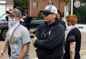 A photo of Ryan Irwin and several other NSC-131 members, at an America Backs the Blue event in Mansfield MA during September 2020. They stand in the northeast corner of the lot for the Mansfield Gulf and Service Station on Chauncy Street, with the buildings across the street visible in the background. In the foreground are Irwin and NSC-131 co-founder Chris Hood. Just behind them are a number of people, including members Anthony Petruccelli and Cameron Anthony. Irwin wears black pants, a gray Smokey the Bear "If Not You, Who?" graphic t-shirt, black plastic-framed rectangular lenses, and a camo Red Sox baseball cap. He wears a white bandana to conceal most of his face and neck. His arms are decorated with a number of tattoos. Both Irwin and Hood appear to be in verbal confrontation with someone off-camera, with Irwin in the midst of gesticulating.