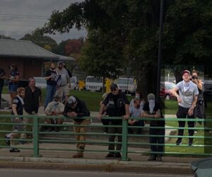 A photo of several NSC-131 members amongst participants at an America Backs the Blue event in Mansfield MA during September 2020. They stand or sit behind the green fencing on the south side of the North Common, with the Common itself and the buildings on Crocker Street visible in the background. Standing partially on the fence is Irwin, apearing ready to throw a Roman salute. Irwin wears black pants, a gray Smokey the Bear "If Not You, Who?" graphic t-shirt, black plastic-framed rectangular lenses, a camo Red Sox baseball cap, and black sneakers with white soles. He wears a white bandana around his neck. His arms are decorated with a number of tattoos, not discernable from a distance. Along with Irwin on his side of the fence are, from left-to-right, NSC-131 members Cameron Anthony, Anthony Petruccelli, Chris Hood, who appears to be Zach Brackett, and another unidentified member.