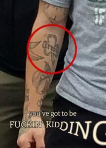 A close-up of Irwin's right arm, cropped from a photo of the America Backs the Blue event in Mansfield MA during September 2020. A tattoo on his inside upper forearm is of a clover with what appears to be the letters "Irwi" printed in it, the last letter disappearing to perspective. Text has been overlaid on the image, saying "you've got to be FUCKIN' KIDDING," with the word "kidding" getting furiously larger and more distorted with every letter, the last "G" getting cut off by the image border.