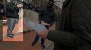 A screenshot of NSC-131's propaganda video of their Portsmouth NH standout against a drag queen story hour during December 2021. Taken outside of the Seacoast Repertory Theatre, a row of members, white men in black clothes and masks, adorned with the number "131" decorating various articles of clothing, stand on a brick sidewalk, holding a banner in the background. A man in the midground hands another man in the foreground a flyer. One man, wearing a black windbreaker and gloves, black sneakers with white soles, gray slacks, a black gaiter with the NSC-131 logo on it in white, and a black baseball cap decorated with a white sonnenrad, holds the edge of the banner. He's removed his right glove to manage a cell phone. The back of his right hand visibly has a black tattoo on it. The banner and flyer have been intentionally redacted, and the man holding his phone is highlighted.