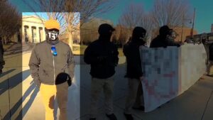 A screenshot of NSC-131's propaganda video of their Boston MA standout against Brigham & Women's Hospital during January 2022. Taken in front of Stoneman Centennial Park on Francis Street, a row of members, white men in black clothes and masks, adorned with the number "131" decorating various articles of clothing, stand on a concrete sidewalk, holding a banner in the foreground. One man, standing next to the banner but not holding it, wears a black windbreaker and gloves, khaki slacks, a black gaiter with the numbers "131" on it in white, a khaki tan baseball cap, and what appear to be corrective lenses with black frames. He wears a gray Mjolnir symbol from his windbreaker's zipper-pull. Next to the the Mjolnir is a set of blue-lensed aviator sunglasses, tucked into the collar of his windbreaker. His hands are in his pants' pockets. The banner has been intentionally redacted, and the man with his hands in his pockets is highlighted.