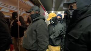 A screenshot of NSC-131's propaganda video of their Boston MA standout against Brigham & Women's Hospital during January 2022. Taken in Haymarket Station, a procession of members, white men in black clothes and masks, adorned with the number "131" decorating various articles of clothing, board an Orange Line train outbound to Oak Grove Station. One man, to the center-right of frame, wears a black windbreaker and gloves, khaki slacks, a black gaiter with the numbers "131" on it in white, a khaki tan baseball cap, and what appear to be corrective lenses with black frames. He wears a gray Mjolnir symbol from his windbreaker's zipper-pull. He wears a set of aviator sunglasses. His head is tilted downward, seeming to watch the gap between the train and the platform. He is intentionally highlighted in the image.