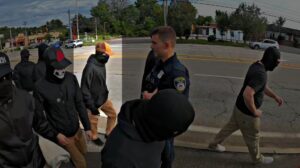 A screenshot of NSC-131's propaganda video of their Framingham MA standout against a migrant shelter during September 2023. Several men in variations of the NSC-131 uniform walk past a Framingham Police officer, on their way to spew invective at a migrant shelter. One member is specifically highlighted. He wears slim-fit khakis, an oversize black hoodie, black sneakers, black gloves with white detailing, a black balaclava, sunglasses, and a khaki trucker cap. He does not make eye contact with the officer as he approaches to pass.