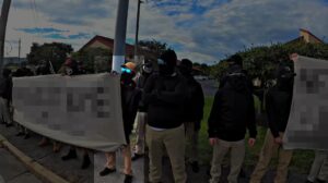 A screenshot of NSC-131's propaganda video of their Framingham MA standout against a migrant shelter during September 2023. Shot with a fisheye effect, disorganized rows of NSC-131 members stand across the way from a local hotel hosting migrants. Two groups of members hold banners, which have been intentionally redacted. In between, a group of members (including NSC-131 member Chase Gilroy) stand unoccupied, looking away from the camera and the shelter. One member, holding the edge of one of the banners and standing next to Gilroy, is specifically highlighted. He wears slim-fit khakis, an oversize black hoodie, black sneakers, black gloves with white detailing, a black balaclava, blue-lensed aviator sunglasses, and a khaki trucker cap. His gaze appears to be trained on the hotel.