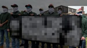 A propaganda photo of NSC-131's Lexington MA standout, as the "New England Minutemen," against a migrant shelter during May 2024. Shot from slightly below and off the the side, a row of men hold an intentionally redacted banner, with their backs turned to the MA National Guard Armory. They almost universally wear blue jeans, green button-up shirts, various colors of baseball cap adorned with the New England Independence flag, several models of sunglasses, and white balaclavas. The shot is framed to grant the illusion of staring boldly into the distance; given the ubiquitous sunglasses, it seems more like they're gormlessly staring at the sun. One man, holding the edge of the banner, is specifically highlighted. He appears to be shorter than most members, some identifiable as usual if unidentified NSC-131 members, who appear to average around 5'10". He also has more obviously sloping shoulders and stubby fingers. His sunglasses are blue-lensed in black, rectangular frames.