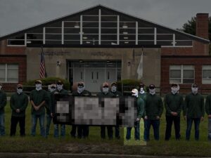 A propaganda photo of NSC-131's Lexington MA standout, as the "New England Minutemen," against a migrant shelter during May 2024. Shot from the sidewalk at Bedford Street, a row of men hold an intentionally redacted banner, with their backs turned to the MA National Guard Armory. They almost universally wear blue jeans, green button-up shirts, various colors of baseball cap adorned with the New England Independence flag, several models of sunglasses, and white balaclavas. One man, holding the edge of the banner, is specifically highlighted. He appears to be shorter than most members, some identifiable as usual if unidentified NSC-131 members, who appear to average around 5'10". He also has more obviously sloping shoulders and stubby fingers. His sunglasses are blue-lensed in black, rectangular frames.